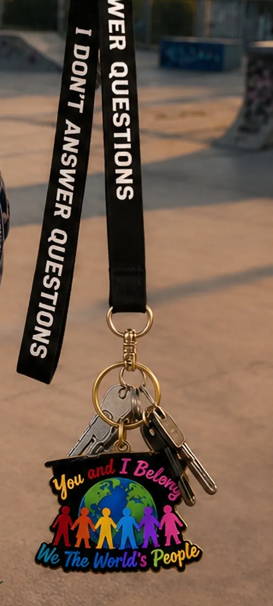 A black lanyard reading "I don't answer questions" attached to a set of house keys and a colorful You and I Belong enamel keychain pendant, hanging outdoors against a blurred sunset background.