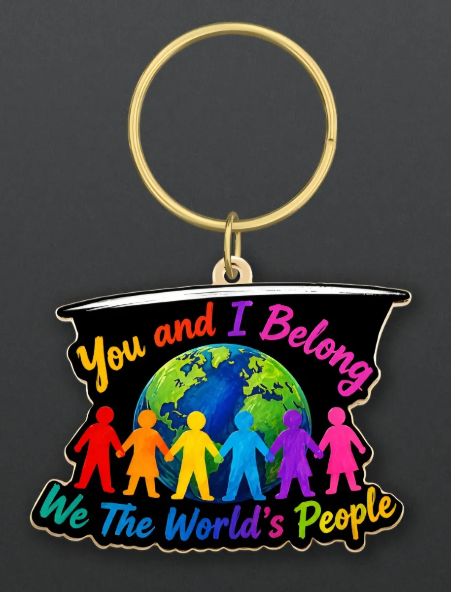 A close-up of the You and I Belong enamel keychain pendant on a dark background. The colorful design features a globe surrounded by diverse figures holding hands, with vibrant text reading "You and I Belong" and "We The World's People