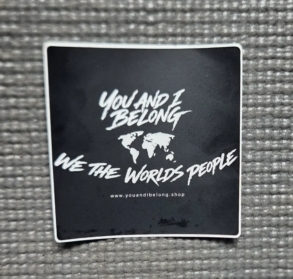 A black square You and I Belong vinyl sticker with a white border, featuring a white world map and the phrase "We The World's People", placed on a grey textured background.