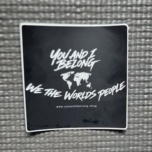 A black square You and I Belong vinyl sticker with a white border, featuring a white world map and the phrase "We The World's People", placed on a grey textured background.
