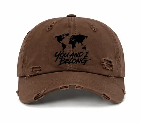 A brown, distressed baseball cap shown from both the front and back. The front features a black world map silhouette and the text "YOU AND I BELONG". The back features the text "WE THE WORLDS PEOPLE" above an adjustable strap.