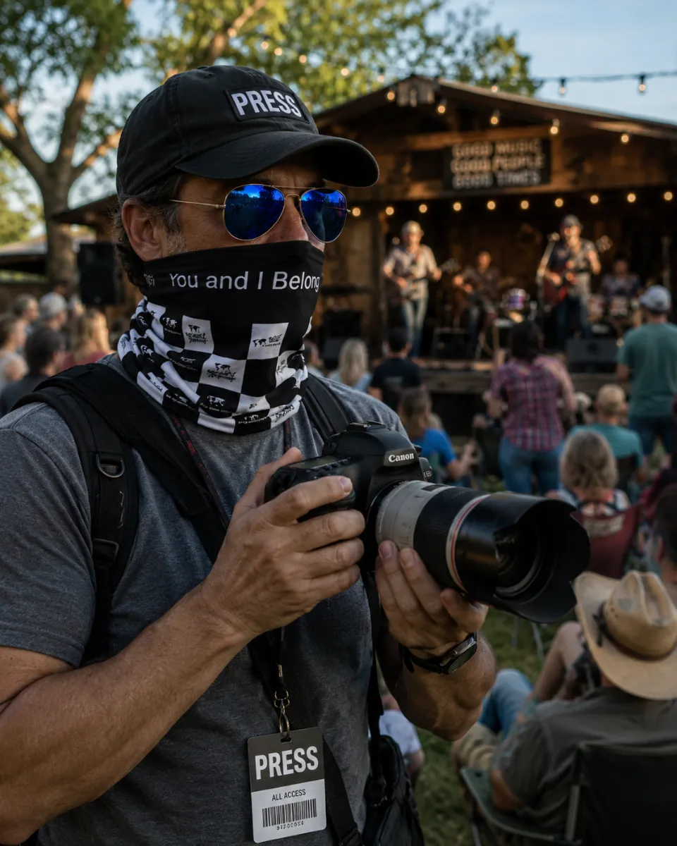 A photographer with a press pass holding a camera at an outdoor concert, wearing sunglasses and a black and white checkered You and I Belong neck gaiter over his face.