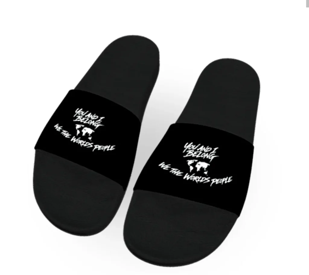 A pair of black slide sandals viewed from above. The straps feature a white world map silhouette with the text "You and I Belong" above it and "We the World's People" below it.