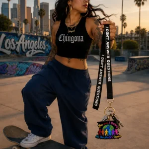 A woman standing with one foot on a skateboard at a skatepark in Los Angeles at sunset, holding a black lanyard that reads "I don't answer questions" attached to keys and a colorful You and I Belong enamel keychain.