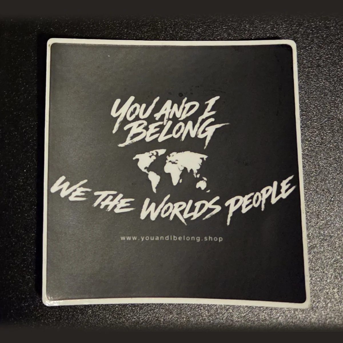 Classic black You and I Belong sticker with a white world map and the words We The Worlds People